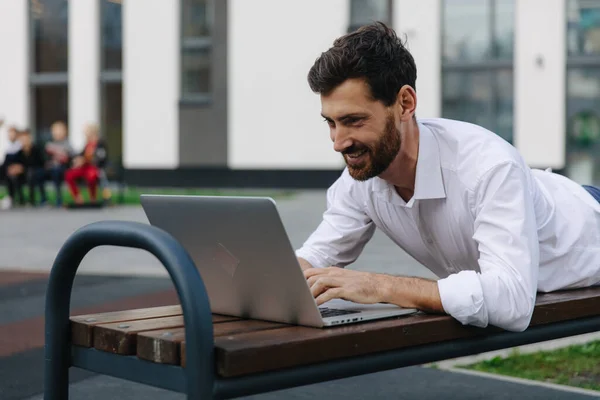 Sonriente hombre acostado en el banco y trabajando en el ordenador portátil — Foto de Stock