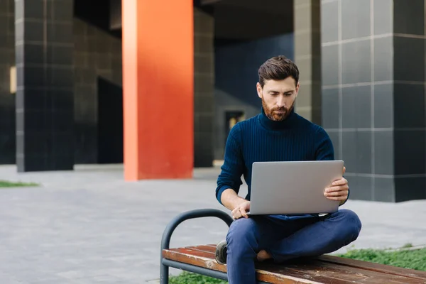 Hombre guapo sentado en el banco y trabajando en el ordenador portátil — Foto de Stock