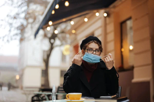 オープンカフェテラスに座っている間に医療用マスクを脱ぐ快適な若い女性 魅力的な女性は屋外で新鮮なホットコーヒーを楽しむ — ストック写真