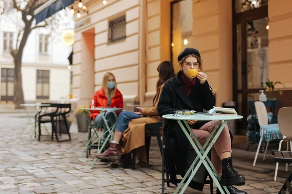 Süslü genç bayan açık kafe terasında tek başına oturuyor ve taze kahve içiyor. Maskeli iki kadının maske takması. Koronavirüsün önlenmesi.