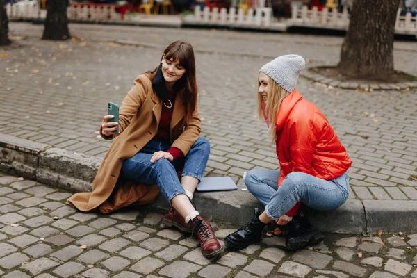 スタイリッシュな若い女性の笑顔と一緒に路上で座ってチャット 幸せな女性は自画撮りのための現代的なスマートフォンを使用して — ストック写真