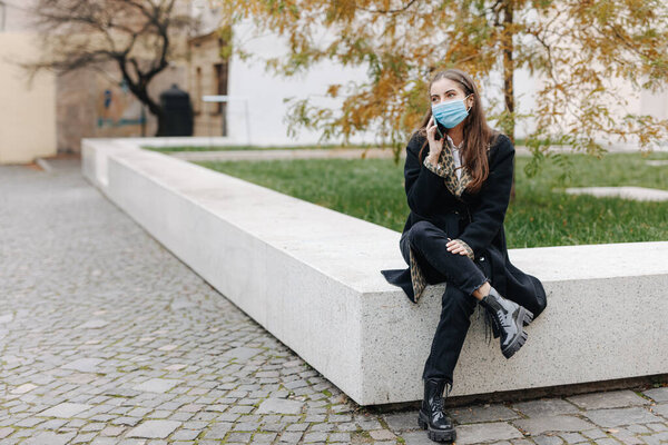 Stylish young woman sitting at city center and using modern mobile for conversation. Female with dark hair wearing medical protective mask during pandemic time.