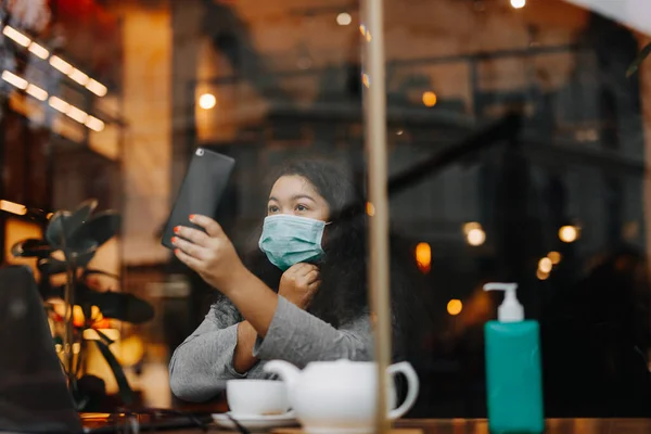 Adorable african lady in medical hygiene mask using laptop and smartphone at cozy cafe. Pretty afro american woman protecting herself from infectious diseases.