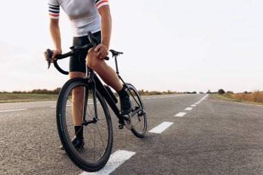 Güçlü vücutlu, bisiklet ve spor ayakkabı giyen, kaldırımlı yolda bisiklete binen genç adama yakın durun. Doğal manzara ve akşam vakti..