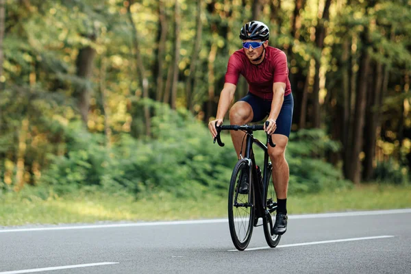 Active young man in helmet and eyewear using black bike for outdoors activity. Bearded cyclist having everyday exercises on paved road.