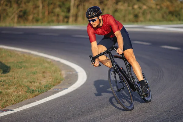 Sporty young man in black helmet, mirrored glasses and activewear riding bike on asphalt road. Bearded cyclist training with high speed outdoors.