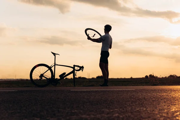 Muscular man in silhouette fixing wheel on broken bike on fresh. Natural landscape and amazing sunset. Sportsman training outdoors.