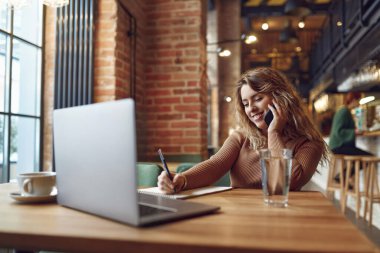 Kıvırcık sarı saçlı gülümseyen kadın kafede kablosuz bilgisayarla oturuyor ve cep telefonuyla konuşuyor. Uzaktan kumanda için modern aletler kullanan serbest çalışan bir kadın..