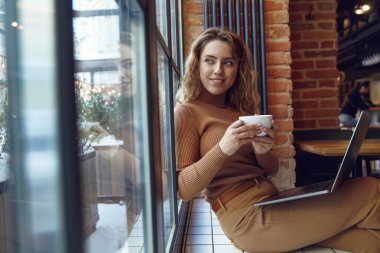 Hoş bir kadın, yerel kafede kablosuz bilgisayarla otururken taze kahve içip pencereye bakıyor. Mutlu bayan boş zamanını sıcak içecek ve modern aletlerle geçiriyor..