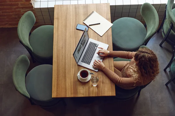 Top view of young woman with blond curly hair sitting at cozy cafe and working on modern laptop. Pretty female doing remote work indoors. Freelance concept.