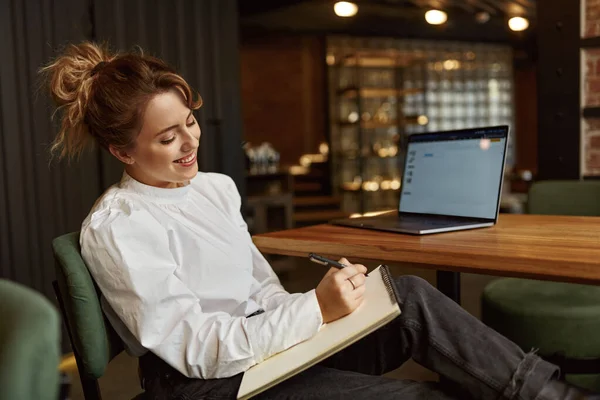 Smiling young woman in white blouse taking notes while working on wireless laptop at cozy cafe. Concept of people, freelance and technology.