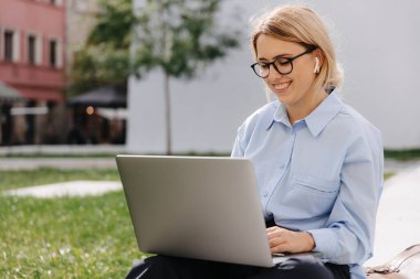 Gözlüklü, sarışın, neşeli genç bayan. İnternette, temiz havada, kablosuz bilgisayarda sörf yapıyor. Modern teknoloji ve şehir yaşam tarzı kavramı.
