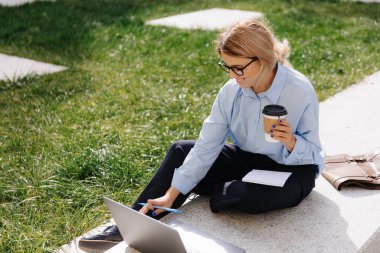 Sarı saçlı güzel genç bir öğrenci modern dizüstü bilgisayarını sokakta ders çalışmak için kullanıyor. Gözlüklü güzel kadın taze kahve içiyor ve notlar yazıyor..