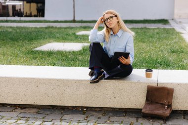 Gözlüklü, elinde dijital tablet olan ve açık havada rüya gören çekici bir sarışın. İnsan kavramı, teknoloji ve modern yaşam tarzı.