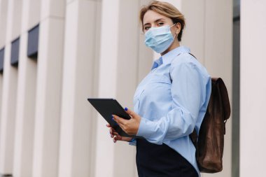 Kahverengi sırt çantalı çekici bir kadın sokakta dikiliyor ve elinde dijital tablet var. Modern aletli genç sarışın dışarıda poz veriyor..