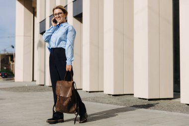 Güneşli havanın tadını çıkaran ve akıllı telefondan konuşan genç bir kadının uzun bir portresi. Sokakta cep telefonu kullanan göz giysili mutlu bir sarışın.