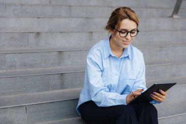 Gözlüklü güzel bir bayan serbest yazar, temiz havada adımlarla yürürken dijital tablet üzerinde çalışıyor. Uzak çalışma ve modern teknoloji kavramı.