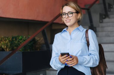 Kahverengi sırt çantalı oldukça çekici bir öğrenci merdivenlerde duruyor ve modern akıllı telefon kullanıyor. Gözlüklü genç kadın dışarıda alet kullanıyor..