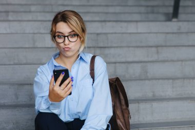 Merdivenlerde otururken modern akıllı telefondan selfie çeken komik sarışın portresi. Aptal suratlı çekici bir kadın açık havada eğleniyor..