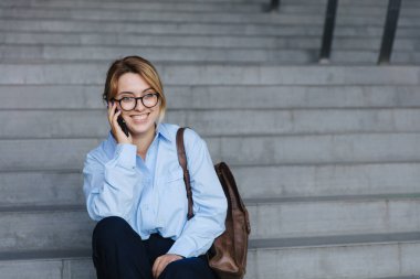 Açık havada merdivenlerde otururken cep telefonunda konuşan neşeli, sarı saçlı genç bir kadın. İnsan kavramı, teknoloji ve iletişim.