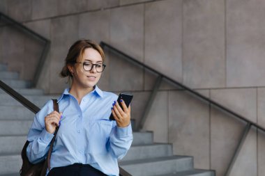 Sıradan giyinen ve gözlüklü güzel bir kız öğrenci merdivenlerde dikilip akıllı telefon kullanıyor. Güzel sarışın temiz havada seyyar ekrana bakıyor..