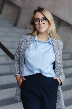 Ofis merkezinin yanında duran resmi kıyafet ve gözlüklerle çekici bir şirket çalışanı. Sarı saçlı genç bayan merdivenlerde poz veriyor..