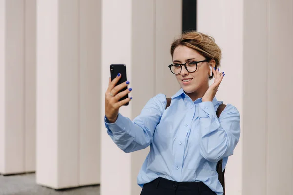 Sarışın, caddede dikilen ve elinde modern akıllı telefon tutan çekici bir genç kadın. Sıradan giyinen çekici bir kadın. Dışarıya video çağrısı yapıyor..