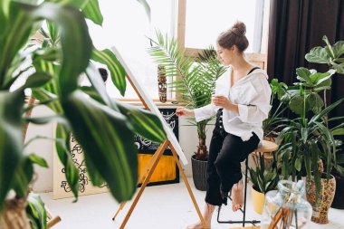 Tuvale fırça ve renk paletiyle resim yapan güzel genç bayanın yan görüntüsü. Çıplak ayaklı, beyaz gömlekli ve siyah pantolonlu pozitif bir sanatçı. Saksıda yeşil bitkiler.