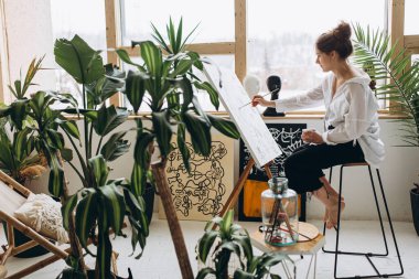 Gündelik giysiler içinde, tuvale renkli resimler çizen çekici kadın ressamın yan görüntüsü. Yalınayak sandalyede oturan ve stüdyoda yaratıcı sürecin tadını çıkaran genç bir kadın..