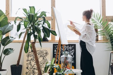 Modern iç mekan ve yeşil bitkiler ile parlak stüdyoda fırçayla resim yapan yaratıcı kadın ressamın yan görüntüsü. İnsan kavramı, hobi ve ilham..