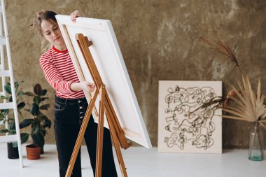 Resim stüdyosunda resim yapmak için resim sehpasını ayarlayan güzel bir genç kadın. Güzel kadın sanatçı çizim için aletler hazırlıyor. İlham konsepti.