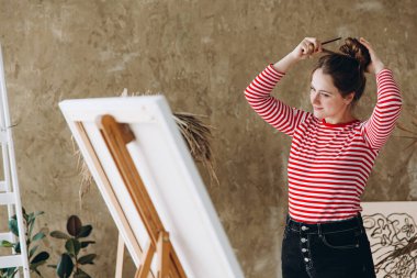 Güzel genç bir kadın resim stüdyosunda sehpanın önünde saçını düzenliyor. Etkilenmiş kadın sanatçı atölyede yaratıcı süreçten zevk alıyor.