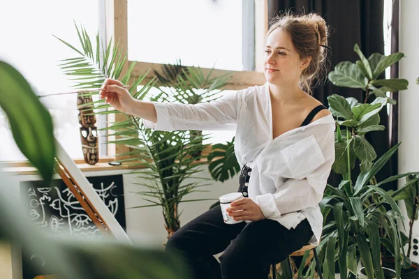 白いシャツを着た愛らしい若い女性と 周りの緑の植物とスタジオでイーゼルに黒いパンツの絵 創造的な趣味と芸術の概念 — ストック写真