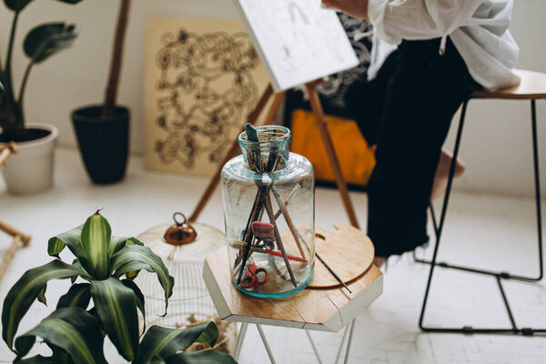 Закрыть стеклянную банку с помощью различных кистей для рисования и цветовой палитры на столе. Размытый фон рисунка художницы на холсте в студии с зелеными растениями вокруг.