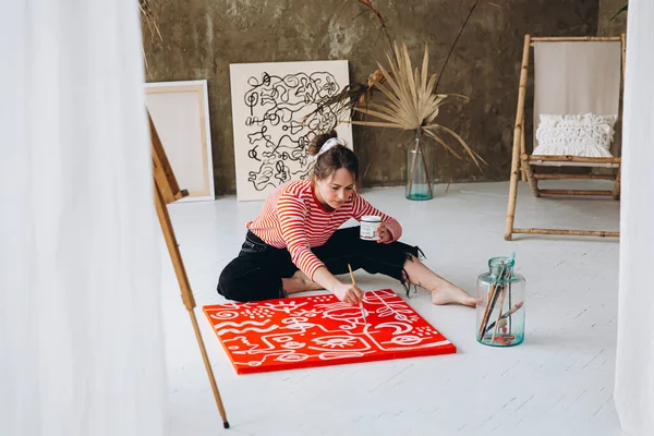 美しい若い女性がアートスタジオで床に座り 赤いキャンバスに白い色で絵を描いています イーゼルに抽象的なパターンを作る女性アーティストにインスピレーションを与えた — ストック写真