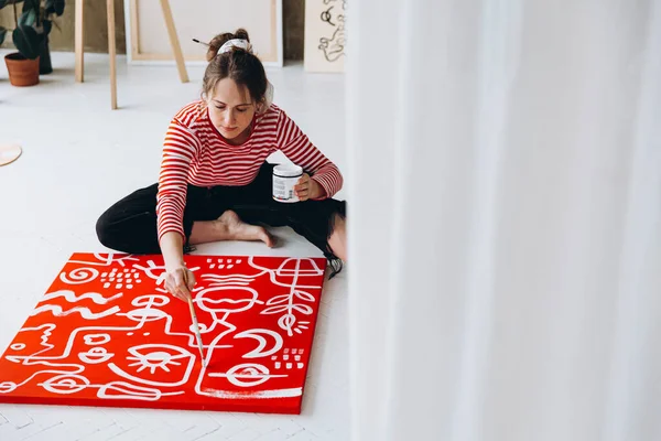 アートスタジオで床に座っている間 赤いキャンバスにカジュアルな衣装の絵で魅力的な若い女性 室内での作業のためにブラシと白の塗料を使用して女性アーティスト — ストック写真