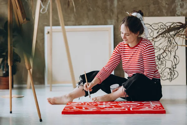 素足で床に座っている間 かなり若い女性が赤いキャンバスに抽象的なパターンを作成します 若い女性アーティストの好きな趣味 — ストック写真