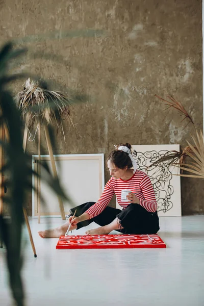 床に座り 赤いキャンバスに絵を描くカジュアルな服装の若い女性にインスピレーションを与えた 抽象的なパターンを描くためにブラシと白の色を使用して女性アーティスト — ストック写真