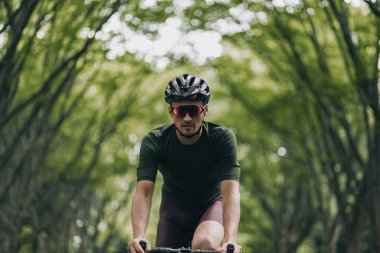 Açık havada tek başına bisiklet süren aktif giysili, kasklı ve gözlüklü genç bir adamın portresi. Yaz parkındaki yeşil ağaçların bulanık arkaplanı. Sağlıklı yaşam tarzları konsepti.