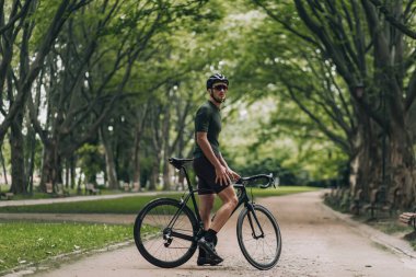 Yeşil şehir parkında siyah bisikletle poz veren atletik vücutlu beyaz bir adam. Kasklı güçlü bir sporcu ve açık hava eğitiminden sonra dinleniyor. Bisiklet kavramı.