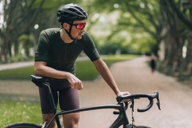 Şehir parkında sabah gezintisi yaparken bisiklete yaslanan genç adamın yan görüntüsü. Spor kıyafetleri, kaskı ve aynalı gözlükleri olan beyaz bir bisikletçi..
