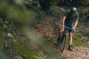 Aktif kıyafetli genç bir adam siyah bir bisiklete binip aşağı bakıyor. Beyaz kasklı sporcu ve gözlüklü açık hava antrenmanından sonra mola veriyor..