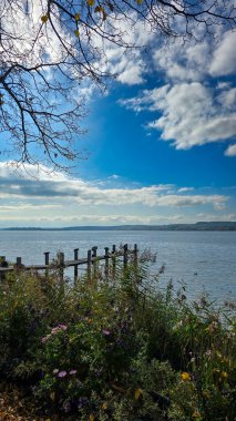 View of the idyllic Lake Constance from the bloom shore on a sunny autumn day. Wooden bridge on the lake. Blue sky with white clouds.