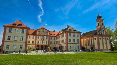 Mainau, çiçek adası, Almanya - 4 Mayıs 2025. Saray Schloss Mainau ve St. Mary Kilisesi Schlosskirche St. Marien yeşil çimenli ve ön planda beyaz çiçekli.