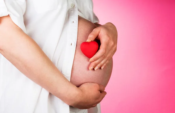 Handen omarmen zwangere buik en hart op een roze — Stockfoto