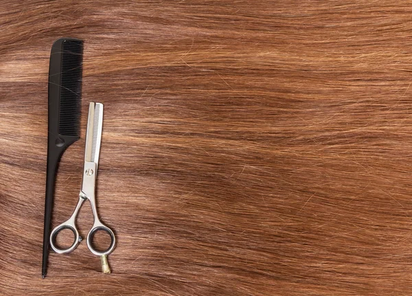 Χτένα και η κομμωτήριο ψαλίδι σε φόντο της όμορφα καστανά μαλλιά. — Φωτογραφία Αρχείου