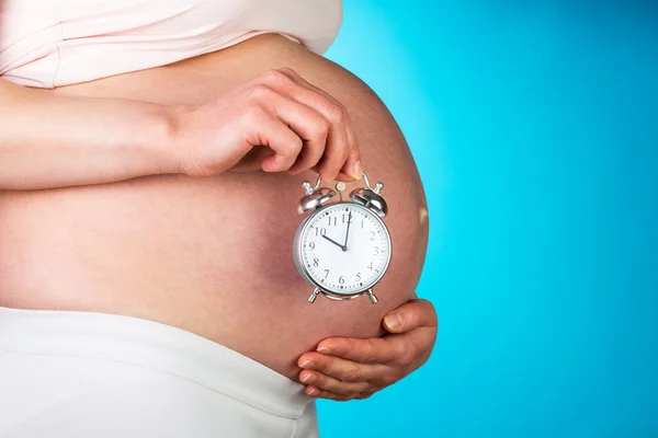 Pregnant woman holding alarm clock on  blue background. — Stock Photo, Image