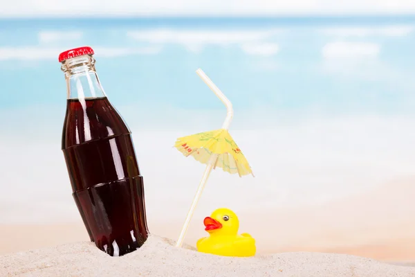 Bouteille de cola, paille et canard en caoutchouc dans le sable contre la mer . — Photo