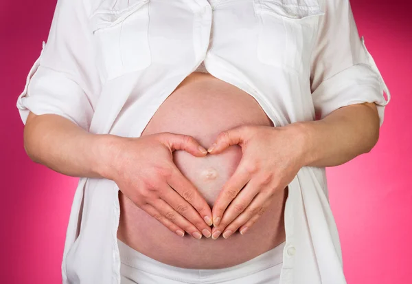 Zwangere vrouw, haar vingers in vorm hart op roze achtergrond. — Stockfoto