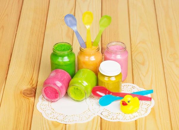 Jars various puree, spoons, rubber duck on background light wood.